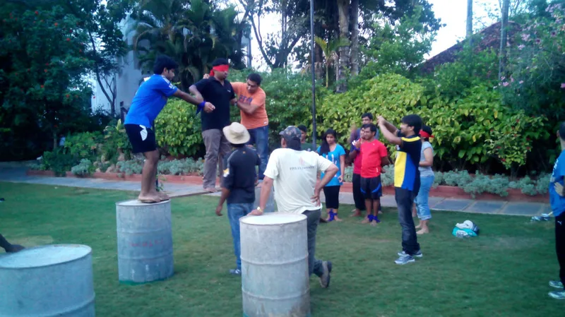 A group of individuals participating in an outdoor team-building activity, balancing on metal barrels and supporting each other, surrounded by greenery.