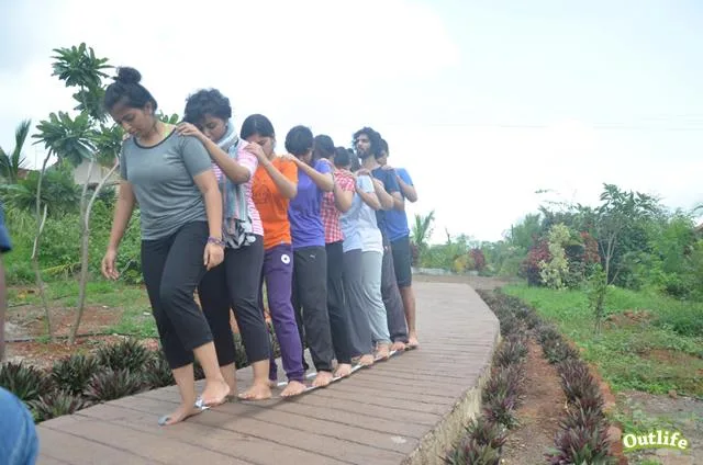 A group of individuals walking in a coordinated line on a narrow pathway, practicing teamwork and balance in an outdoor setting surrounded by greenery.