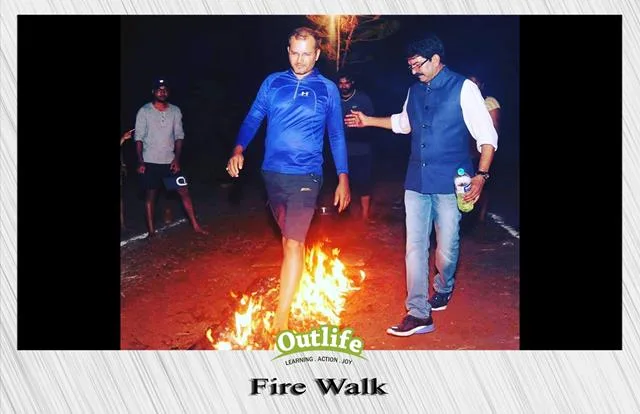 A participant confidently walking barefoot over a bed of burning embers during a fire-walking activity, guided by a facilitator, under the banner of Outlife.