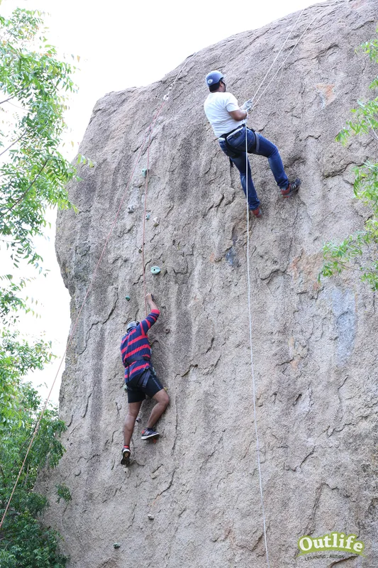 Two people rock climbing on an outdoor natural rock surface with safety ropes, surrounded by green trees