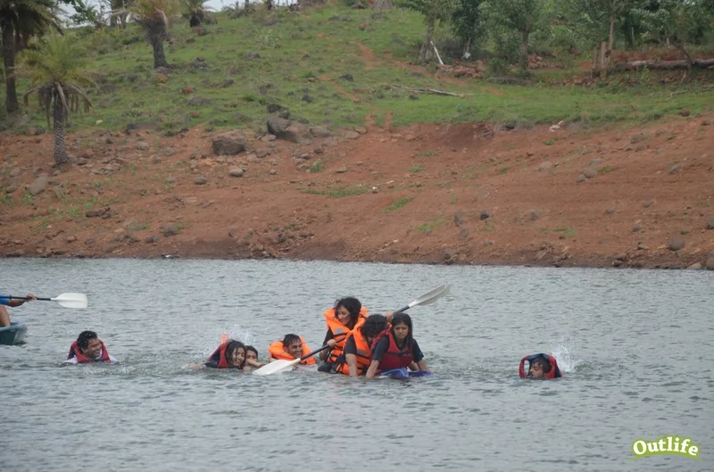 A group of individuals in life jackets engaging in a water activity, with some people in the water near a kayak, set against a backdrop of greenery and a muddy shoreline